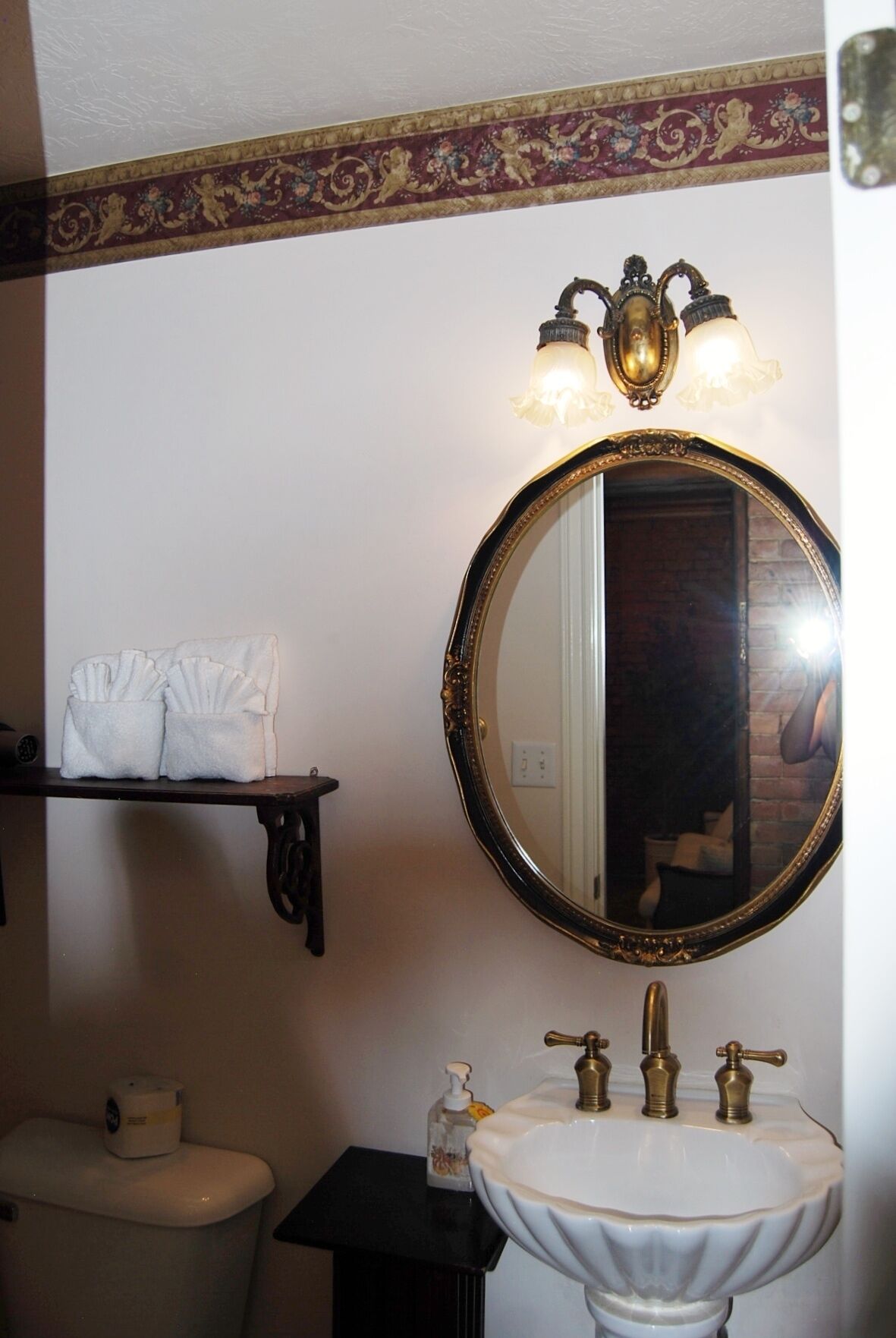 Une salle de bain avec un miroir rond, un lavabo blanc avec des robinets en laiton, une étagère avec des serviettes et des toilettes.