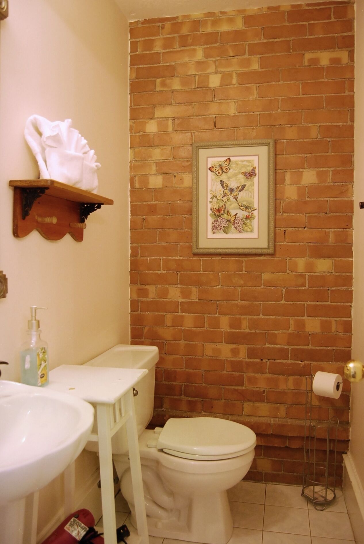 A bathroom with a brick wall, a toilet, a sink, a shelf with a towel, and a framed picture.