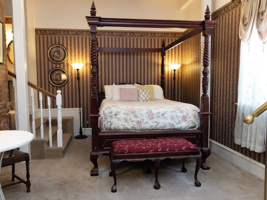 A four-poster bed with a floral bedspread and a matching bench, a white round table, and a staircase with a white railing.