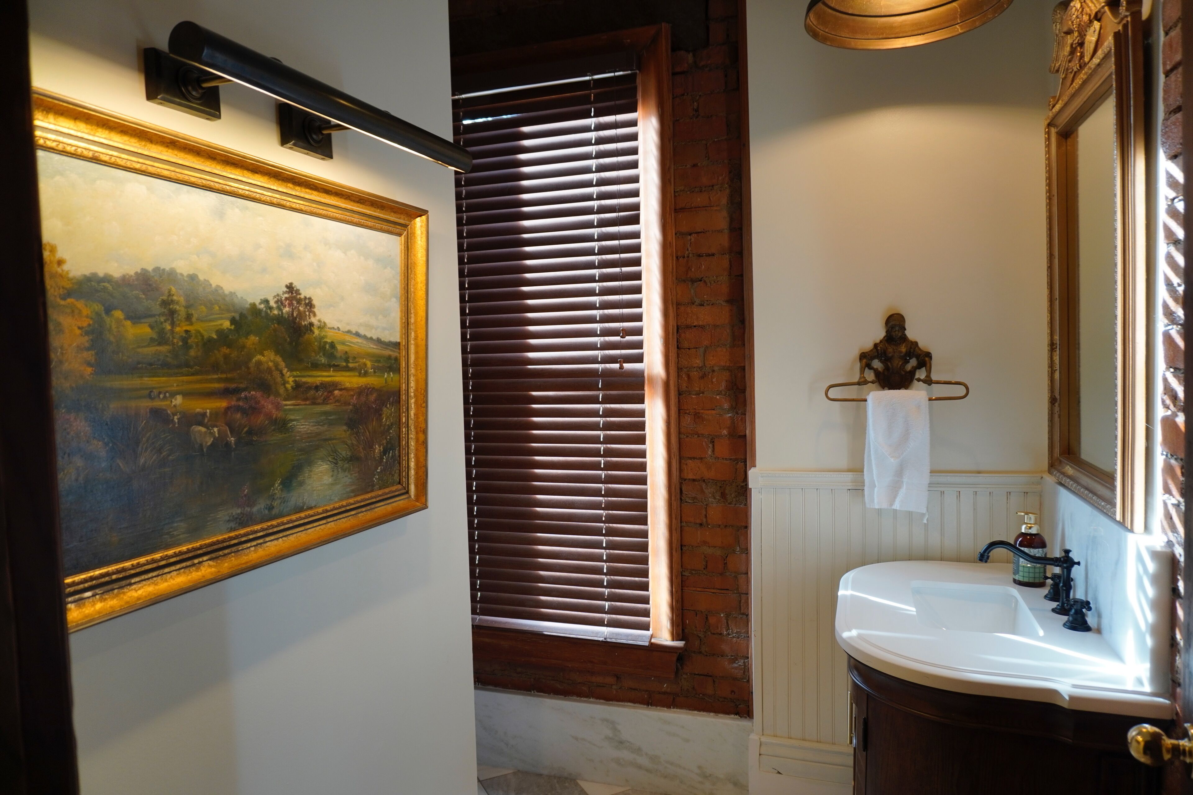A bathroom with a white sink, a mirror, and a framed landscape painting on the wall.
