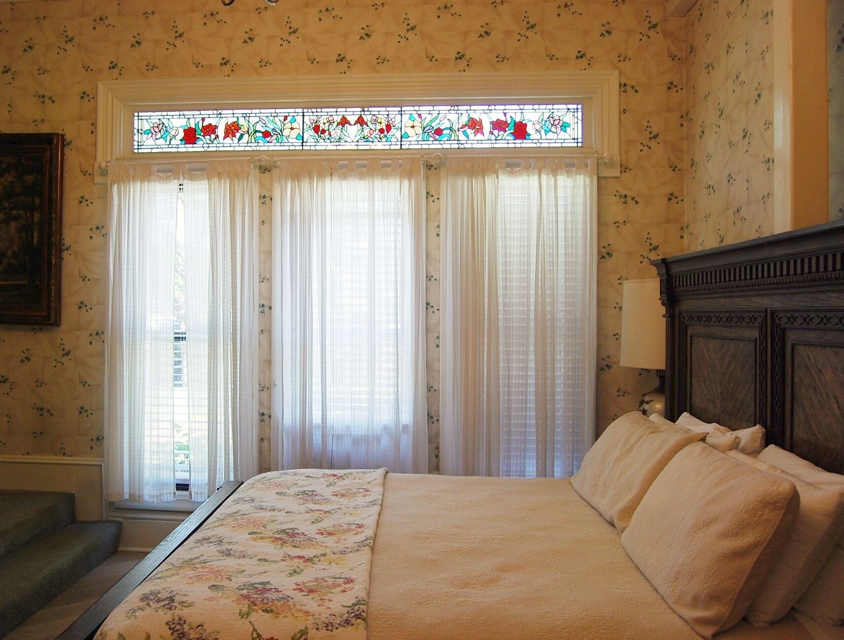 Een slaapkamer met een sprei met bloemenmotief, een houten hoofdbord en een glas-in-loodraam boven de gordijnen.