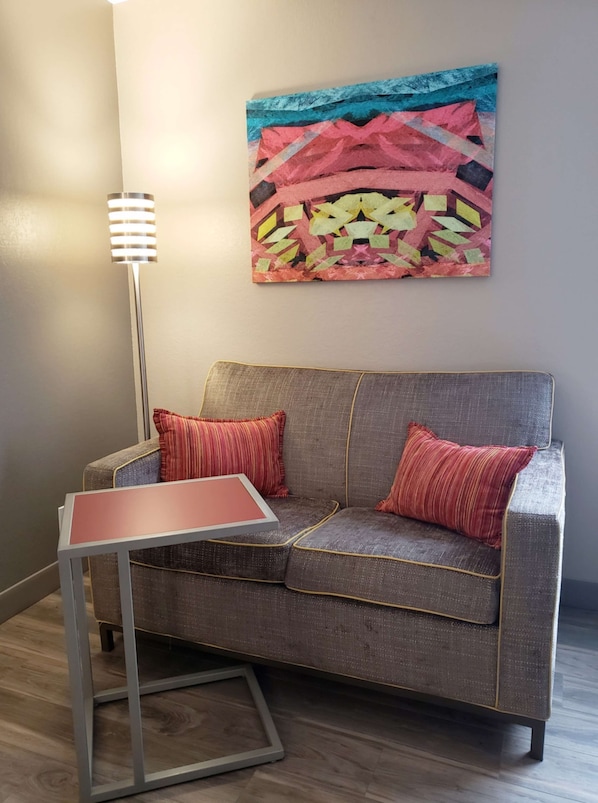 A modern living room with a grey sofa, two red pillows, a small table, and a floor lamp. A colorful abstract painting is on the wall. - Hampton Inn Johnstown (Johnstown)