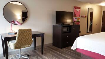 Una habitación de hotel con cama, escritorio y silla, televisor, microondas, espejo y puerta.
