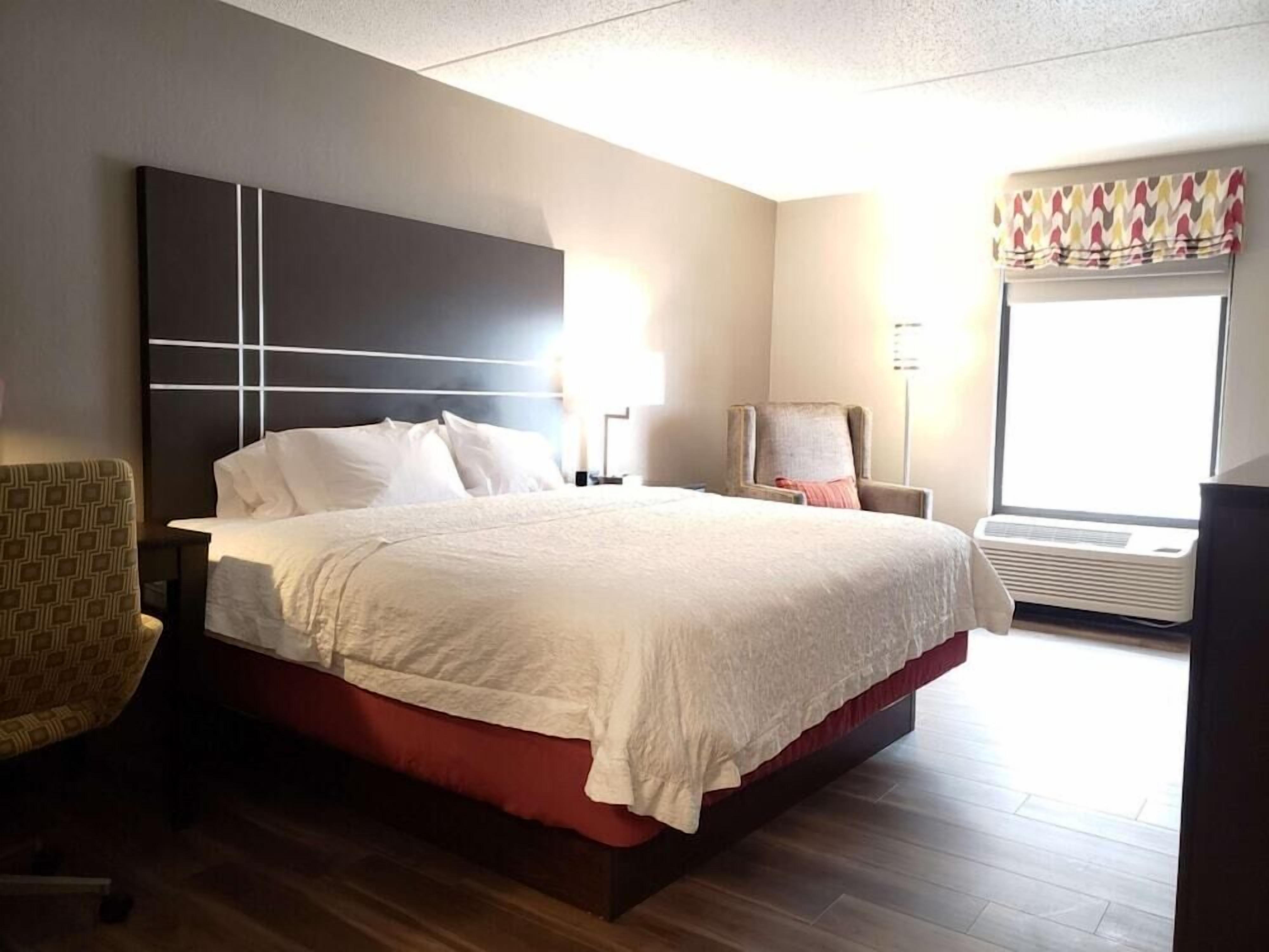 Camera d'albergo con un letto grande, una sedia, un comodino, una lampada e una finestra con condizionatore.