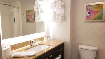 A modern bathroom with a granite countertop, a white sink, a toilet, and a mirror. There are towels stacked on a shelf and a framed artwork on the wall.