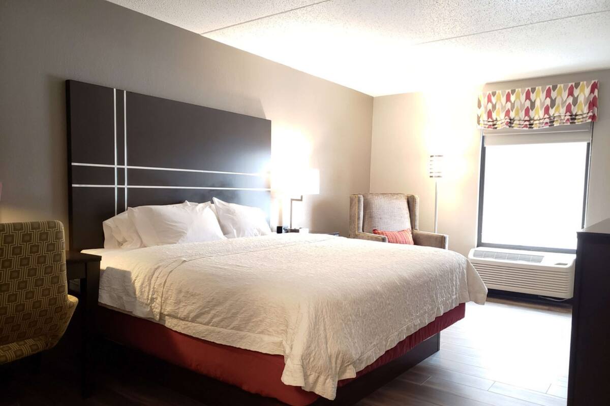 A hotel room with a large bed, a chair, a desk, and a window with an air conditioner.