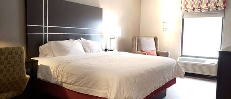 Habitación de hotel con una cama grande, una silla, un escritorio y un ventanal con aire acondicionado.