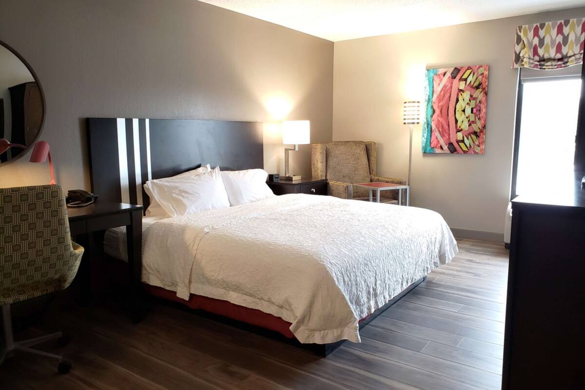 A hotel room with a large bed, a desk, a chair, a lamp, and a painting on the wall.