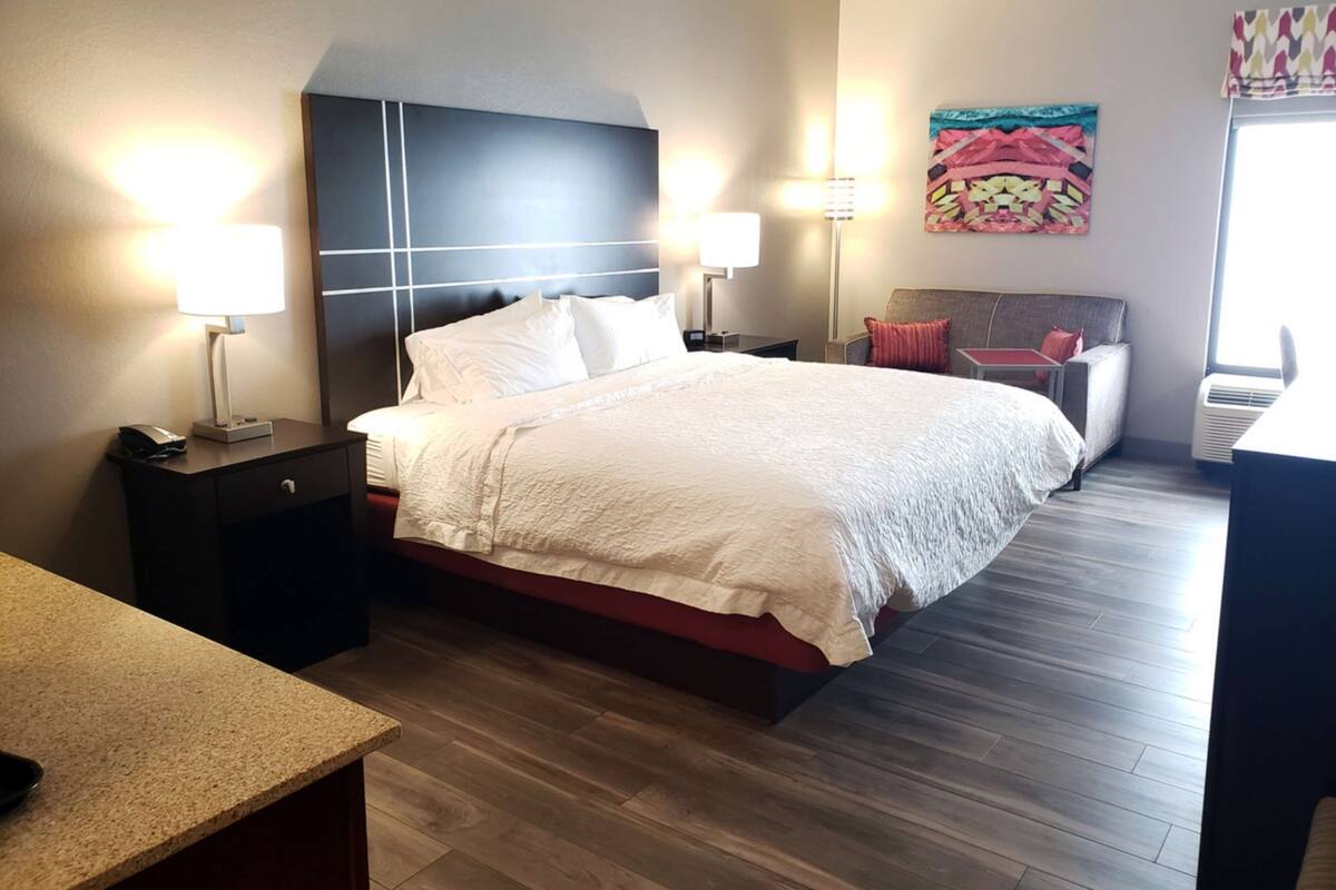 A modern hotel room with a large bed, two bedside tables with lamps, a small desk, and a colorful tapestry on the wall.