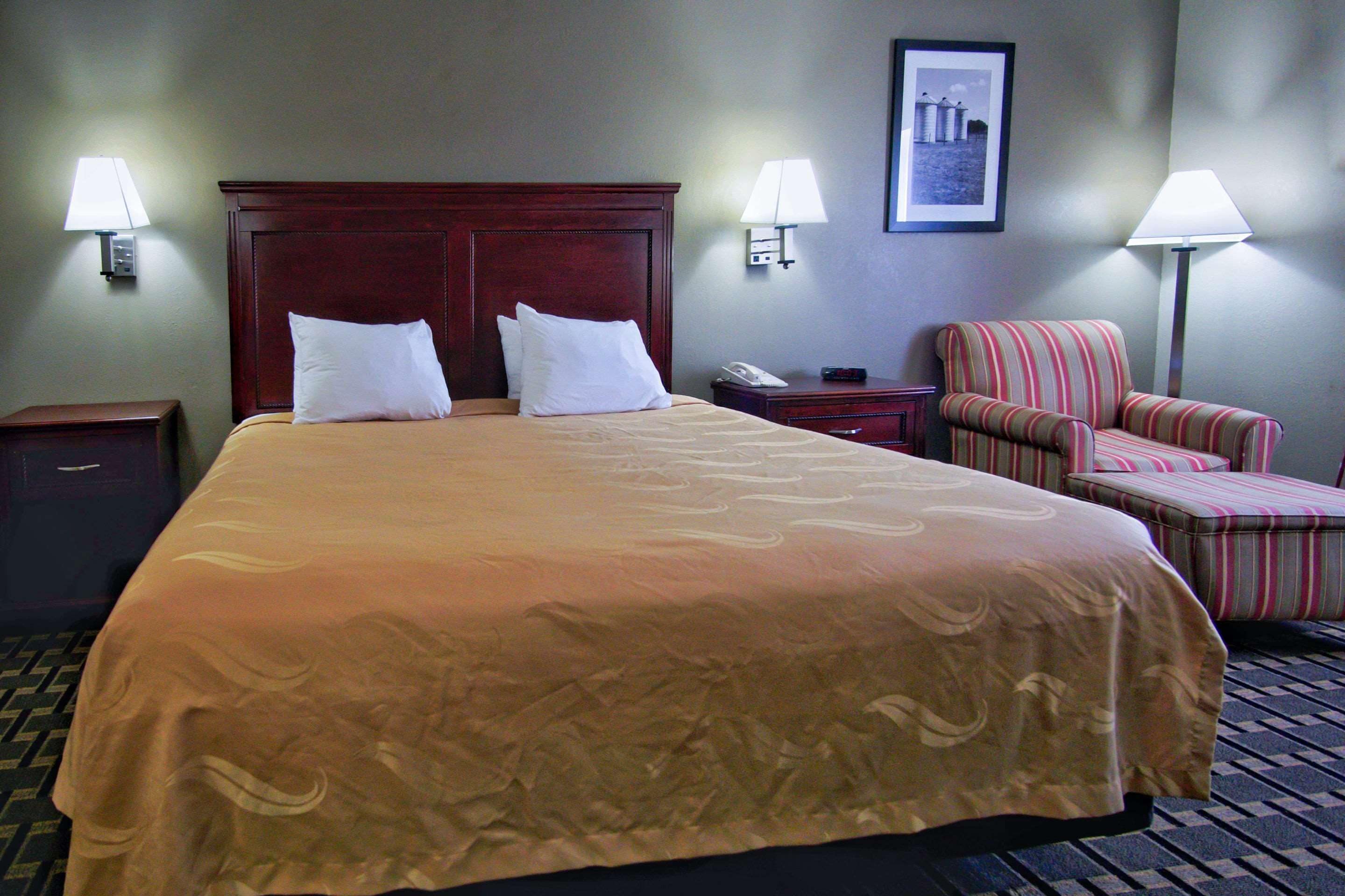 Una habitación de hotel con una cama grande, dos mesitas de noche, una silla y una lámpara.