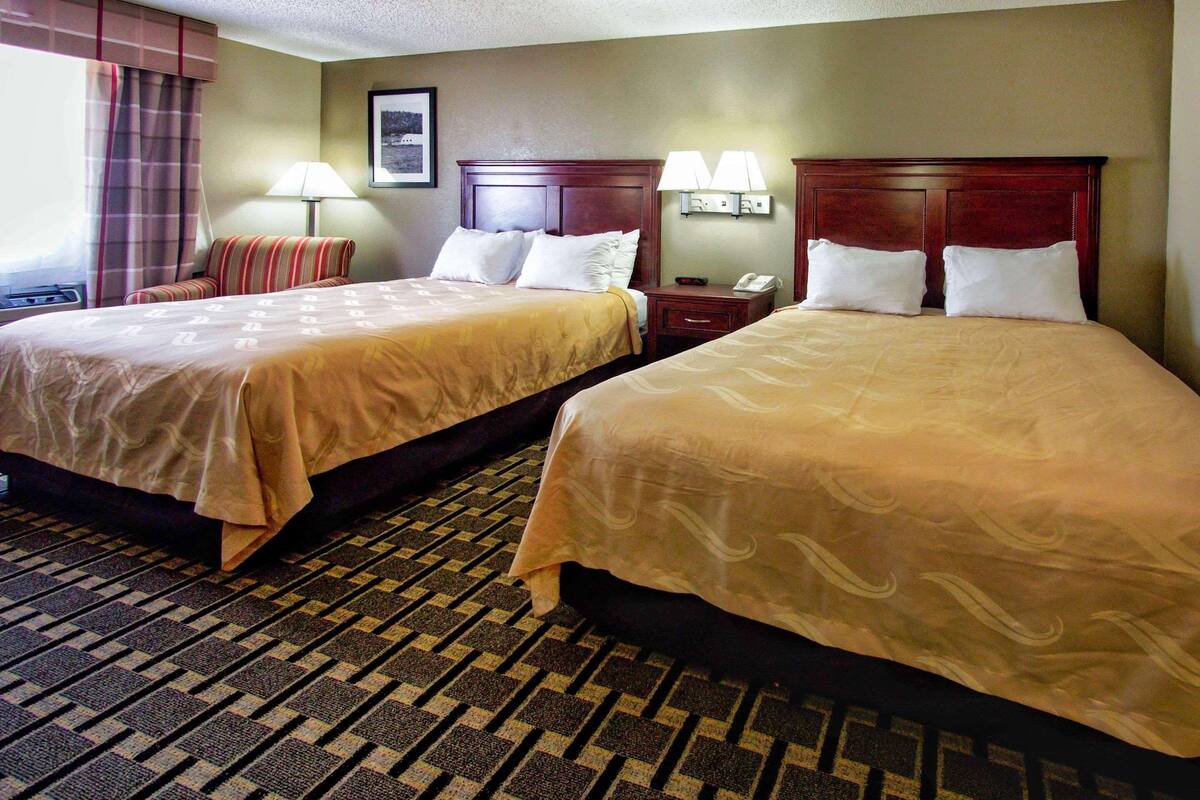 A hotel room with two beds, a carpeted floor, a nightstand with a lamp, and a window with curtains.