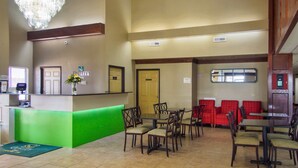 A hotel lobby with a reception desk, green counter, multiple dining tables and chairs, red seating area, and a chandelier.