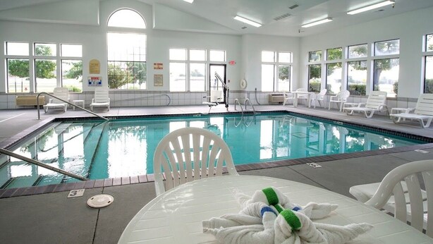 An indoor swimming pool with clear water, white lounge chairs, and a table with towels.
