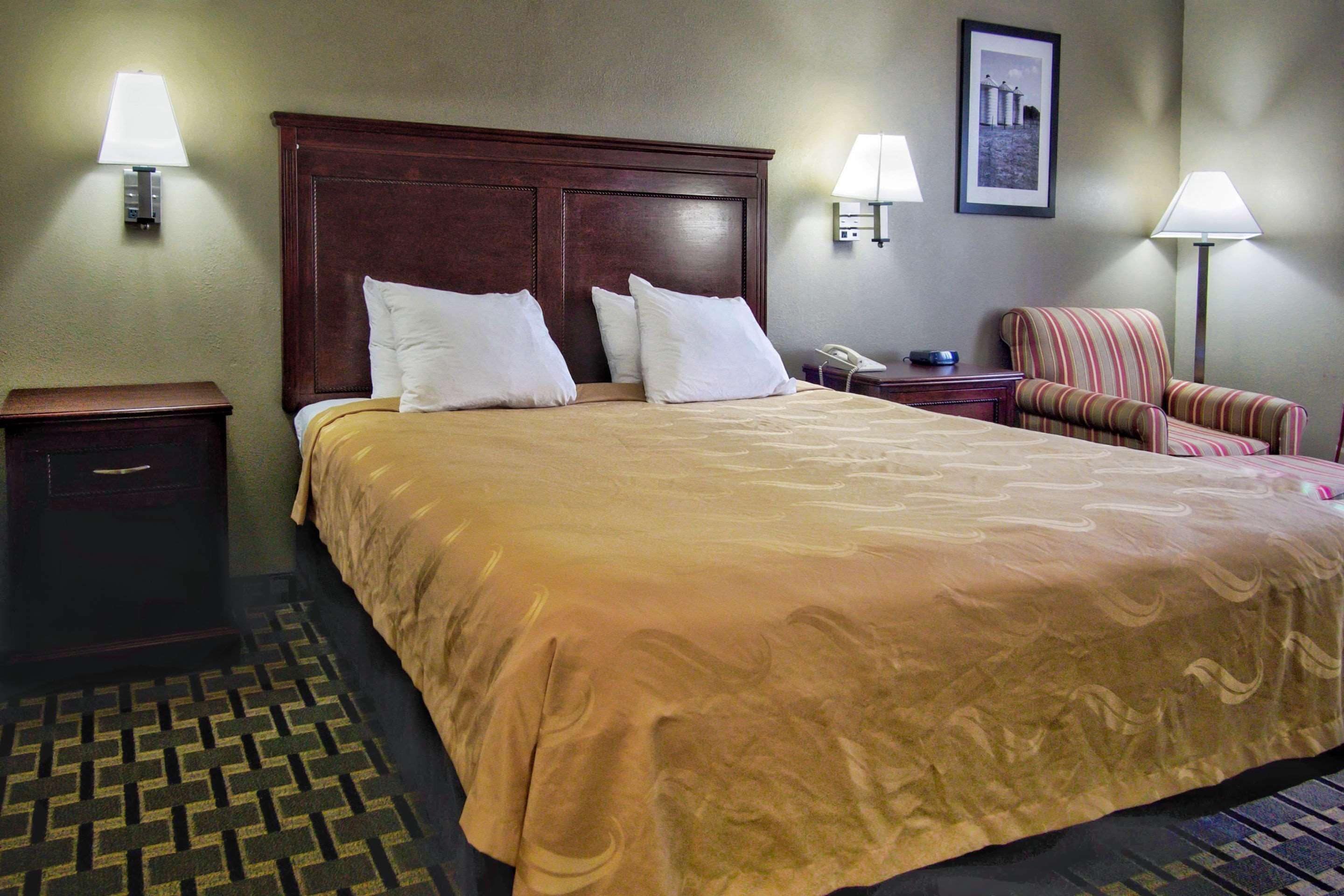 Una habitación de hotel con una cama grande, dos lámparas de noche, una mesita de noche, una silla y un cuadro en la pared.