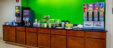 A breakfast bar with a green wall, coffee machines, and cereal dispensers.