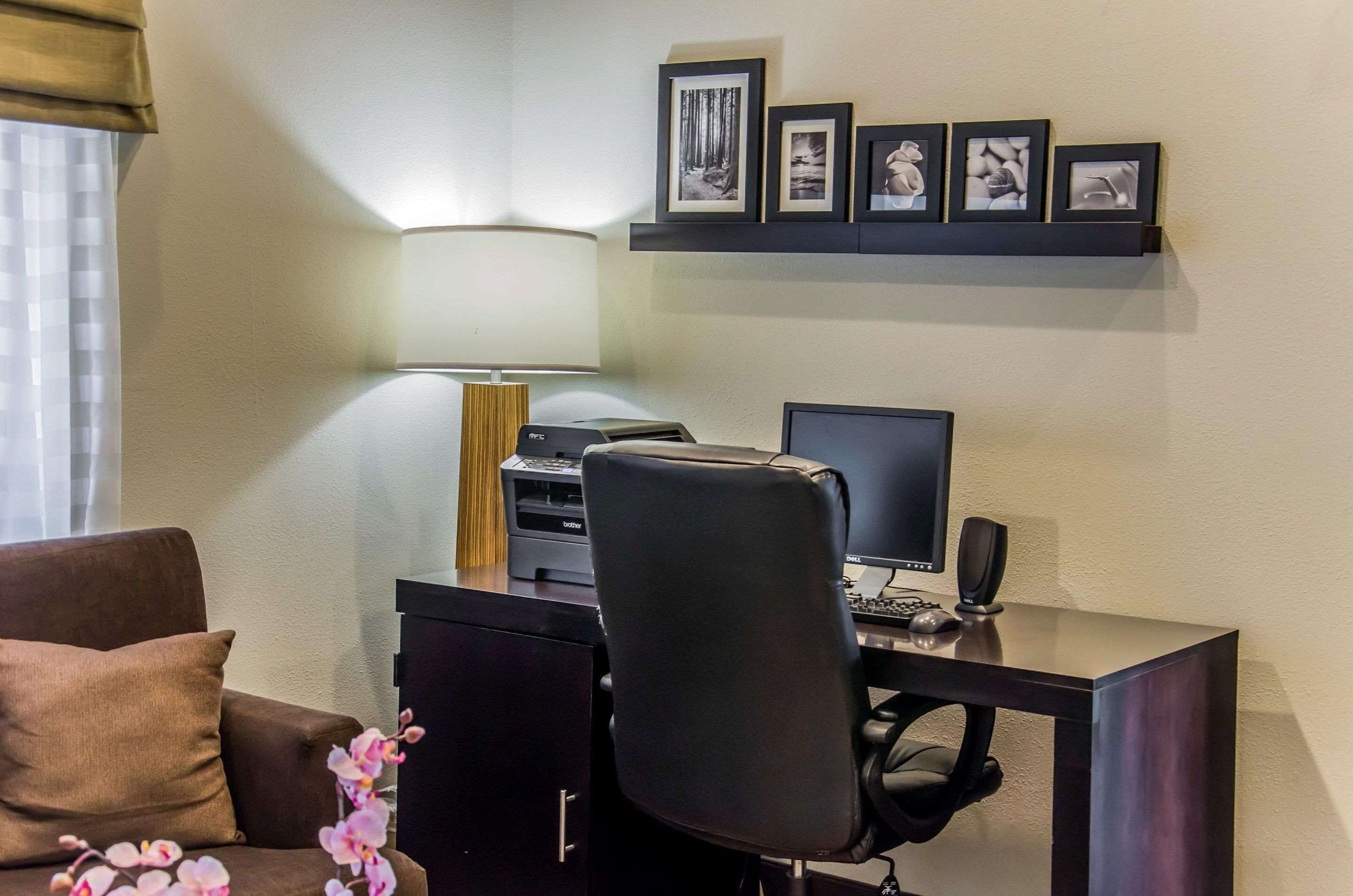 Habitación de hotel con escritorio, computadora, impresora y lámpara. Hay un sofá con un cojín y una mesita con flores.