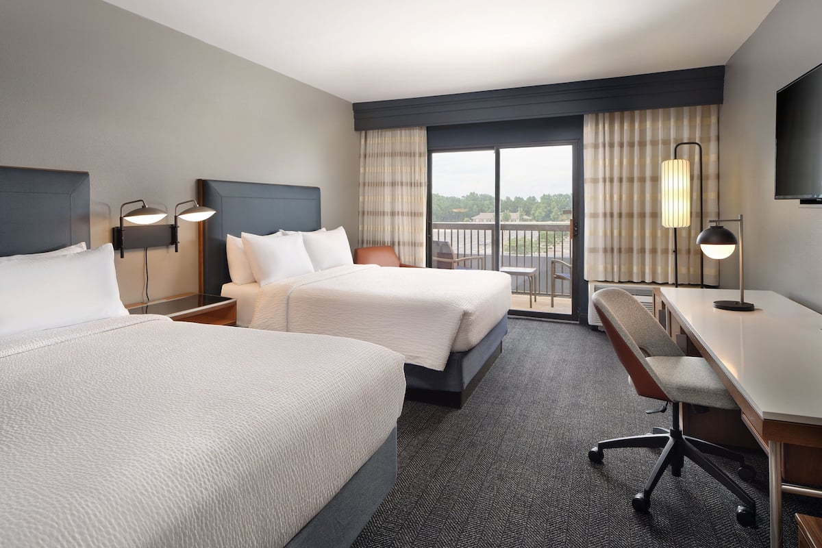 Quarto de hotel com duas camas, uma escrivaninha, uma cadeira e uma varanda com vista.