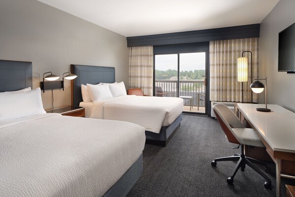 A hotel room with two beds, a desk, a chair, and a balcony with a view.