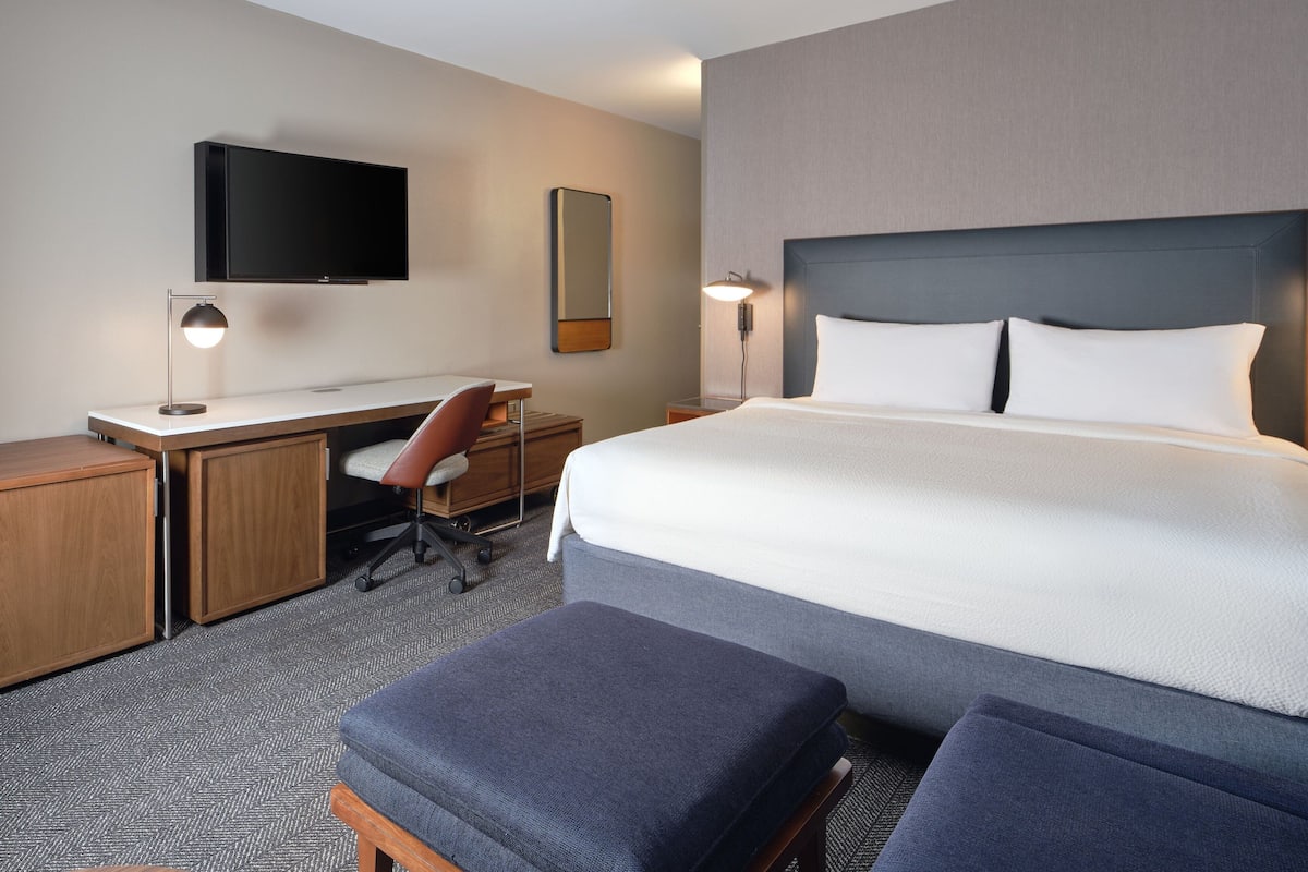 Quarto de hotel com uma cama grande, uma escrivaninha com cadeira, televisão e espelho.