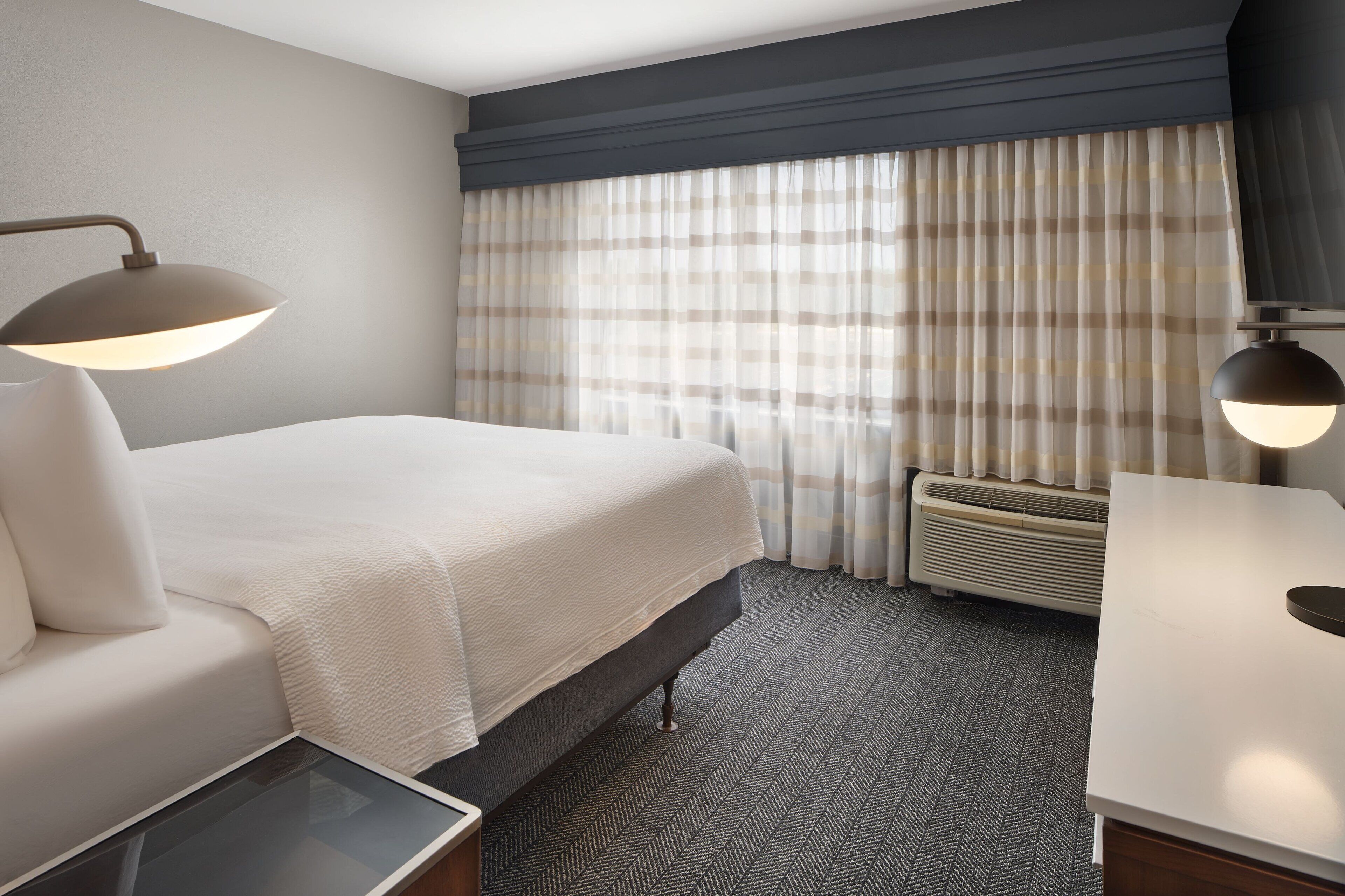 A hotel room with a bed, bedside table, lamp, air conditioning unit, and a view of the outdoors through curtains.