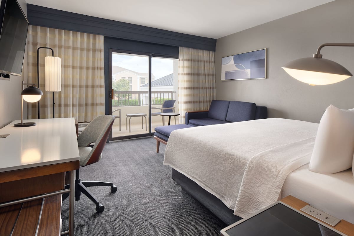 Quarto de hotel com cama, escrivaninha, cadeira, televisão e varanda com vista para casas.