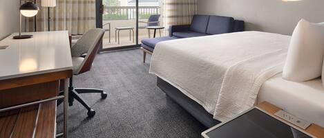 A hotel room with a bed, a desk, a chair, a TV, and a balcony with a view of houses.