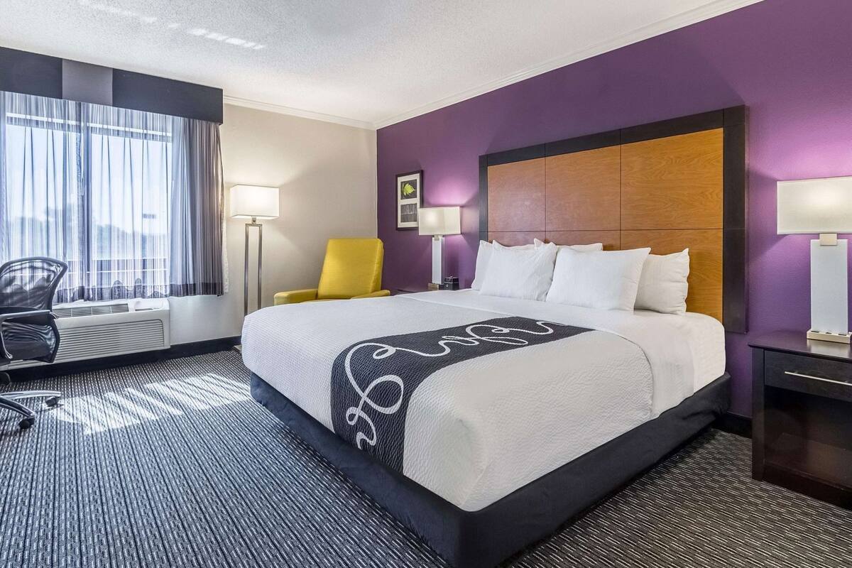 Una habitación de hotel con una cama grande, una silla, un escritorio y un ventanal con cortinas.