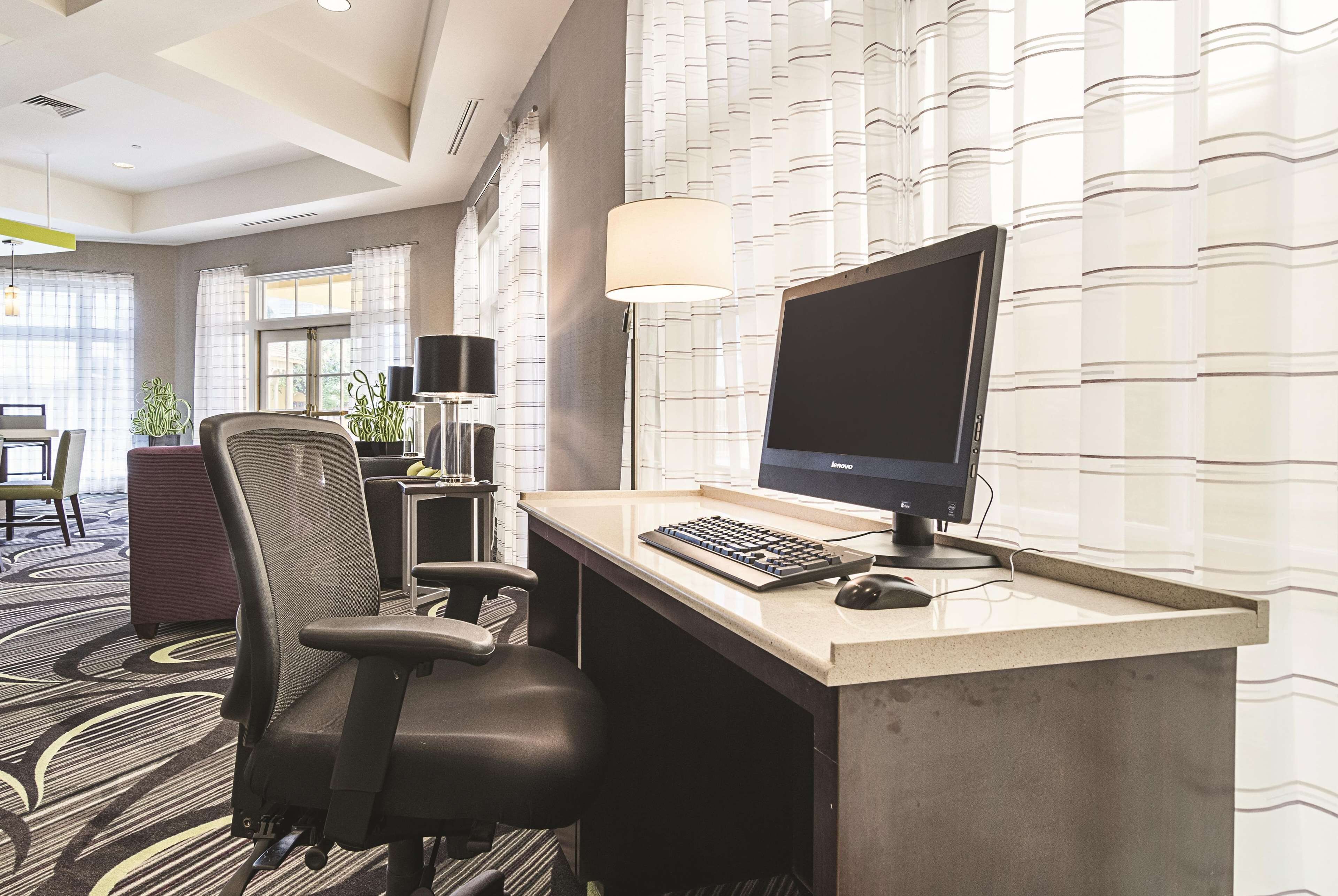A hotel room with a desk, computer, and office chair. There is a lamp, a potted plant, and a window with curtains.