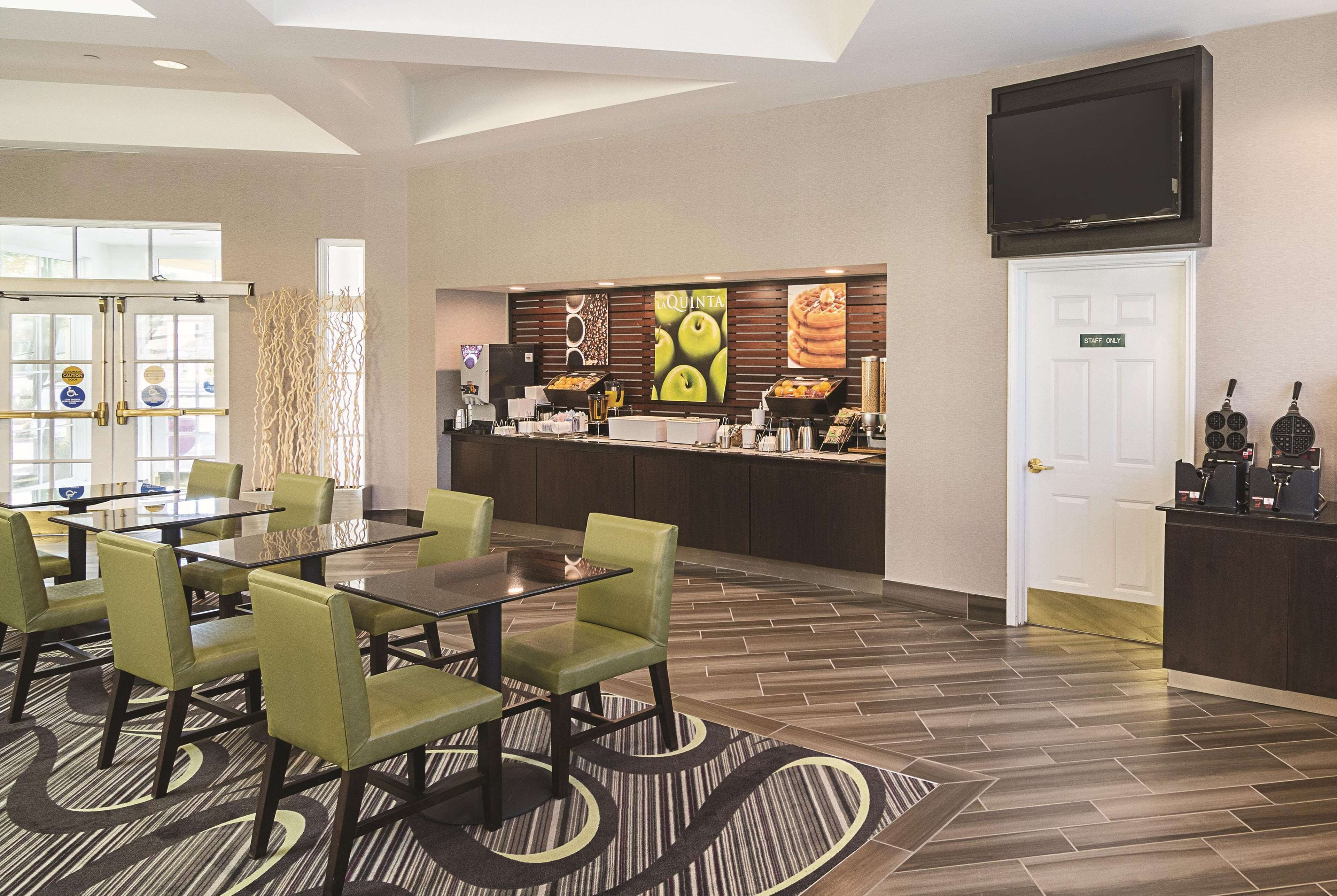 A hotel lobby with a reception desk, a dining area with green chairs and a glass table, and a wall with food display.