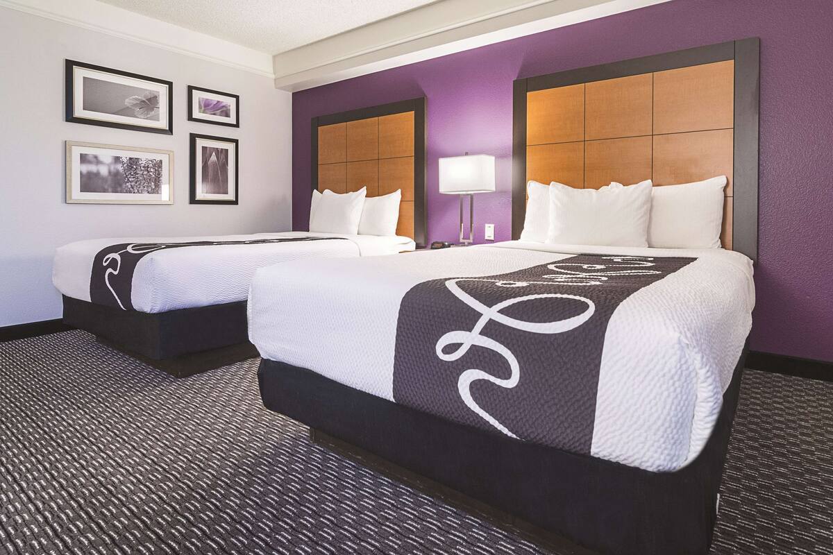 Habitación de hotel con dos camas, cabecera con diseño de panel, lámpara de pared y cuadros en la pared.