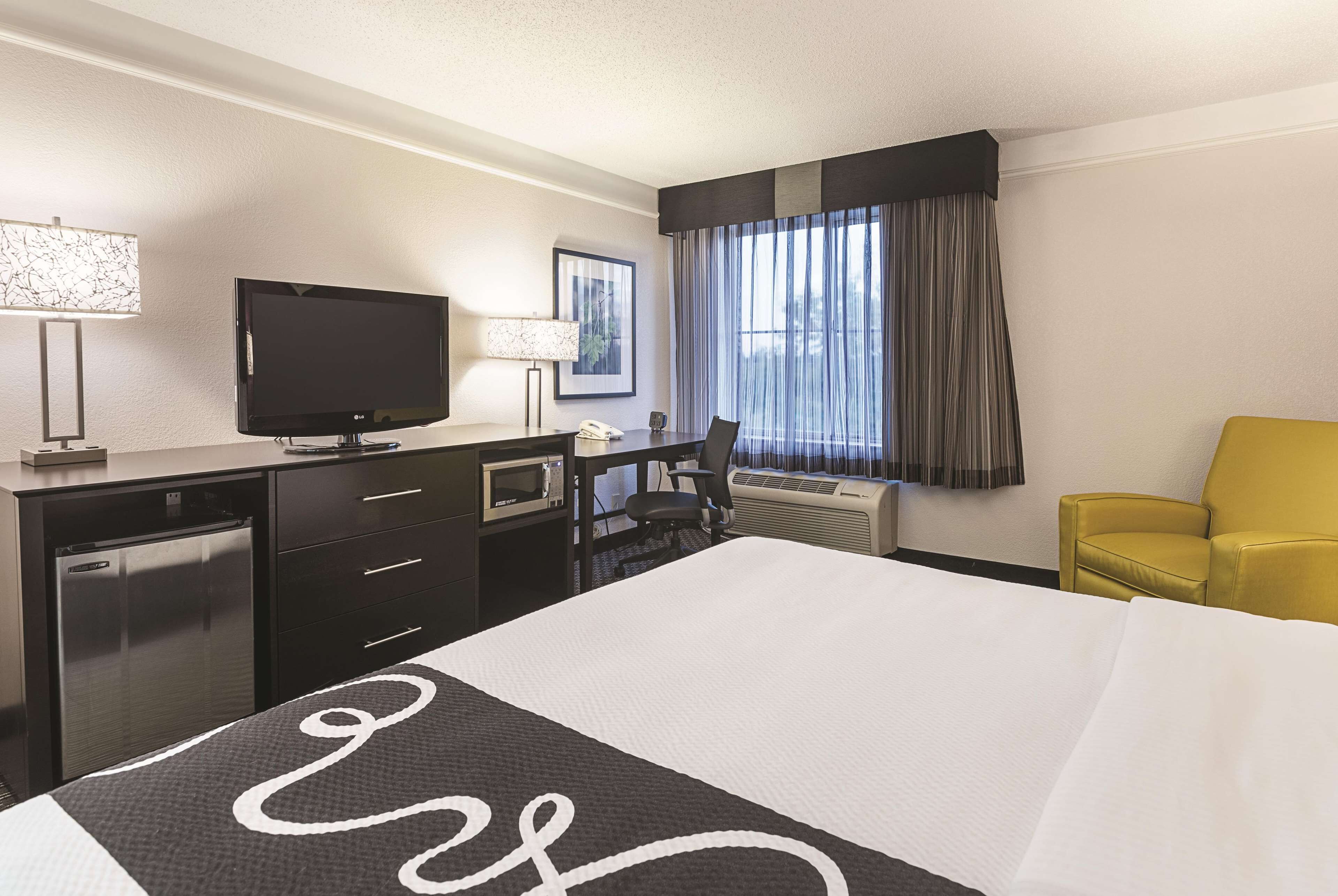 Habitación de hotel con cama, escritorio con televisor, silla, lámpara y ventana con cortinas.