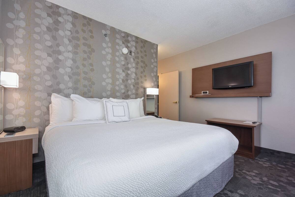 A hotel room with a large bed, a TV mounted on the wall, and a nightstand with a lamp.