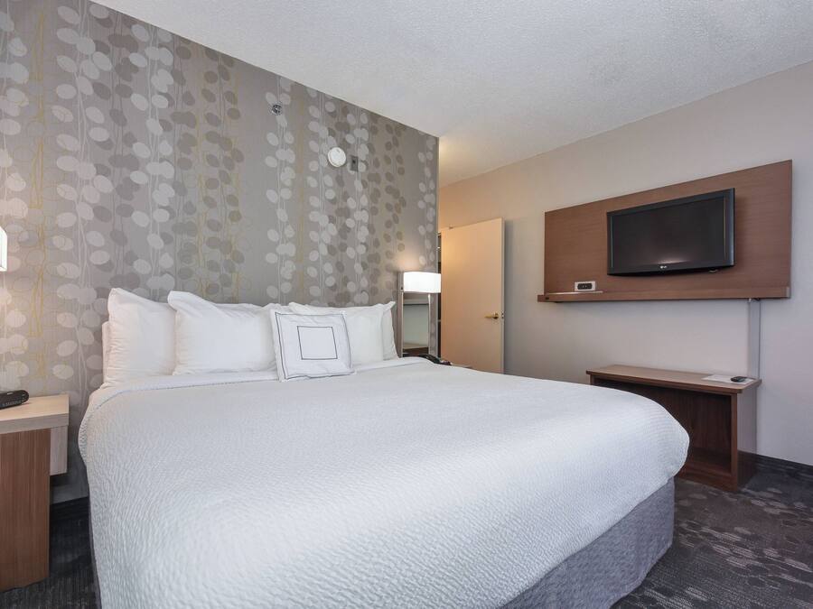 Una habitación de hotel con una cama grande, un televisor montado en la pared y una mesita de noche con una lámpara.