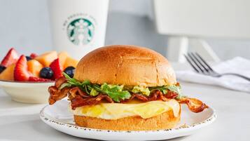 Un sandwich du petit-déjeuner sur une assiette, avec une tasse Starbucks et un bol de fruits en arrière-plan.
