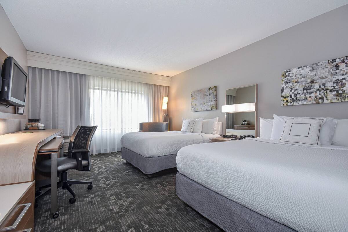 A hotel room with two beds, a desk, a chair, a television, and artwork on the walls.