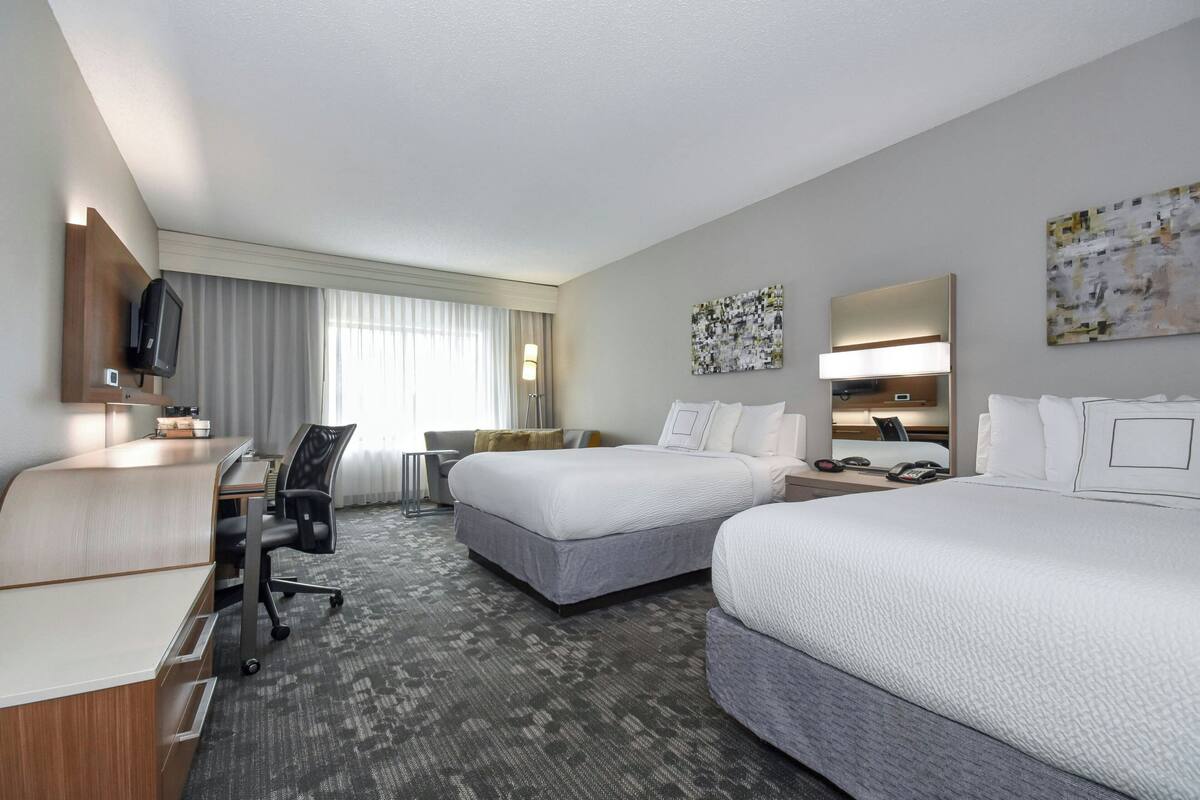 A hotel room with two beds, a desk, a chair, a TV, and artwork on the walls.