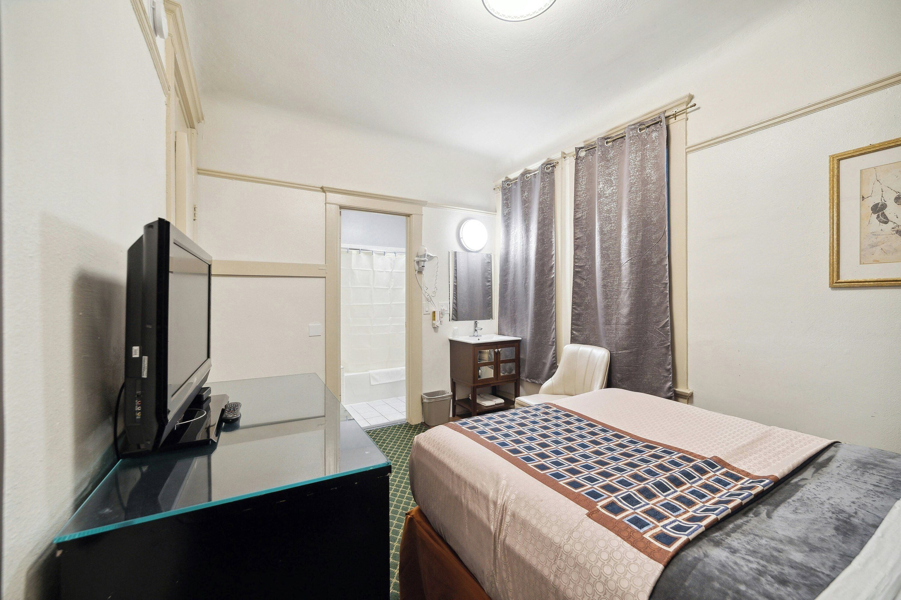 A hotel room with a bed, a TV, a desk with a computer, a bathroom with a shower, and a mirror.