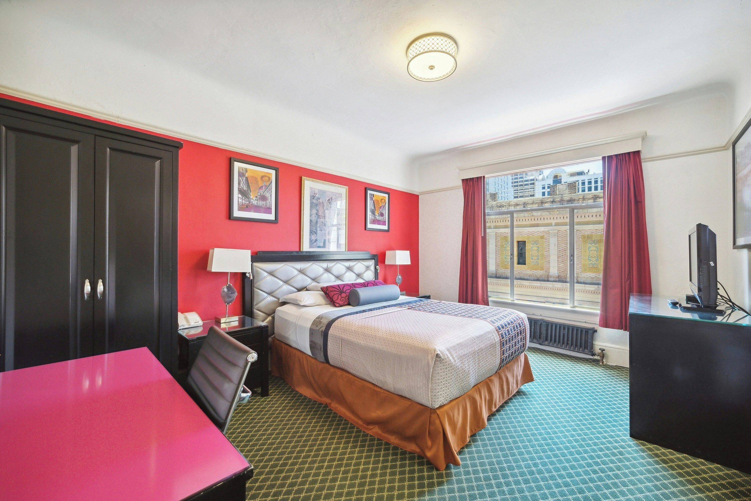 A hotel room with a bed, a desk, a chair, a window with a view of buildings, and a red wall.