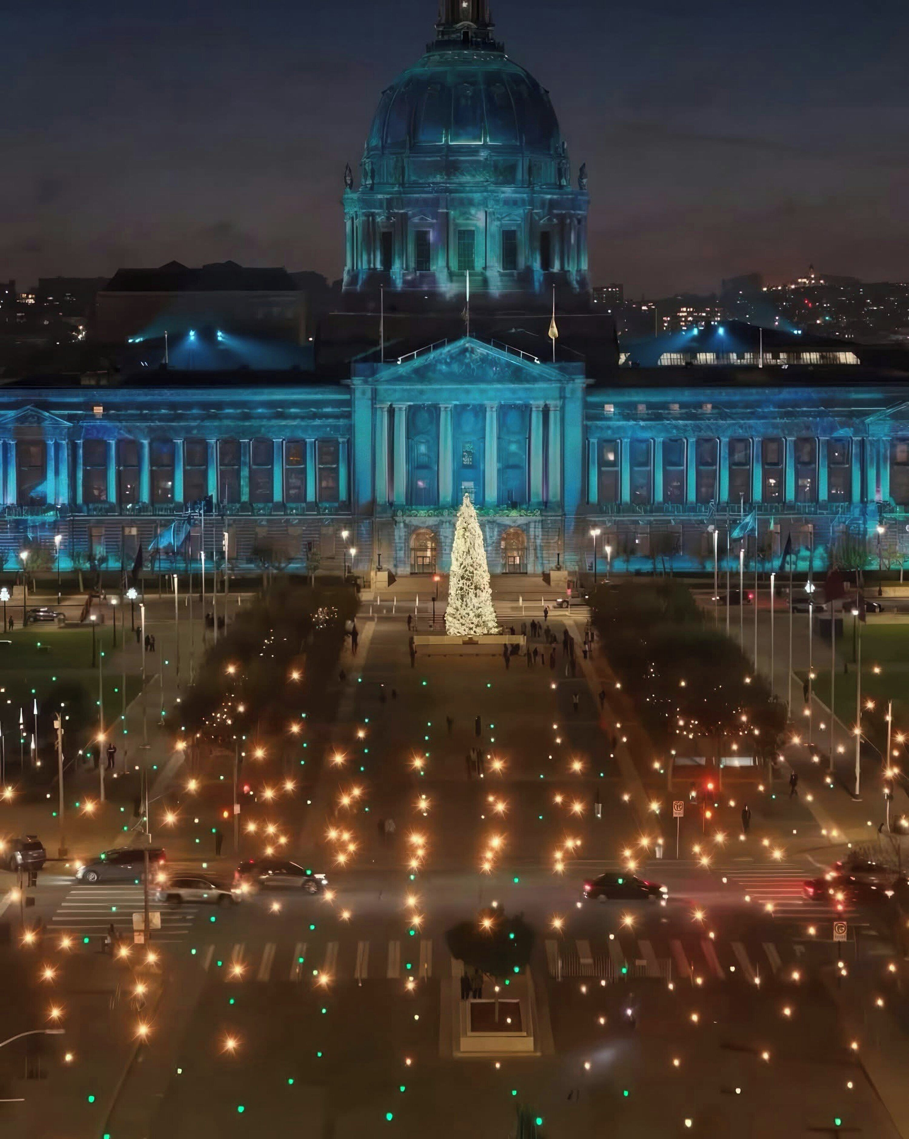 A large building with a prominent dome and columns, a central tree decorated with lights, and a well-lit plaza with street lamps.