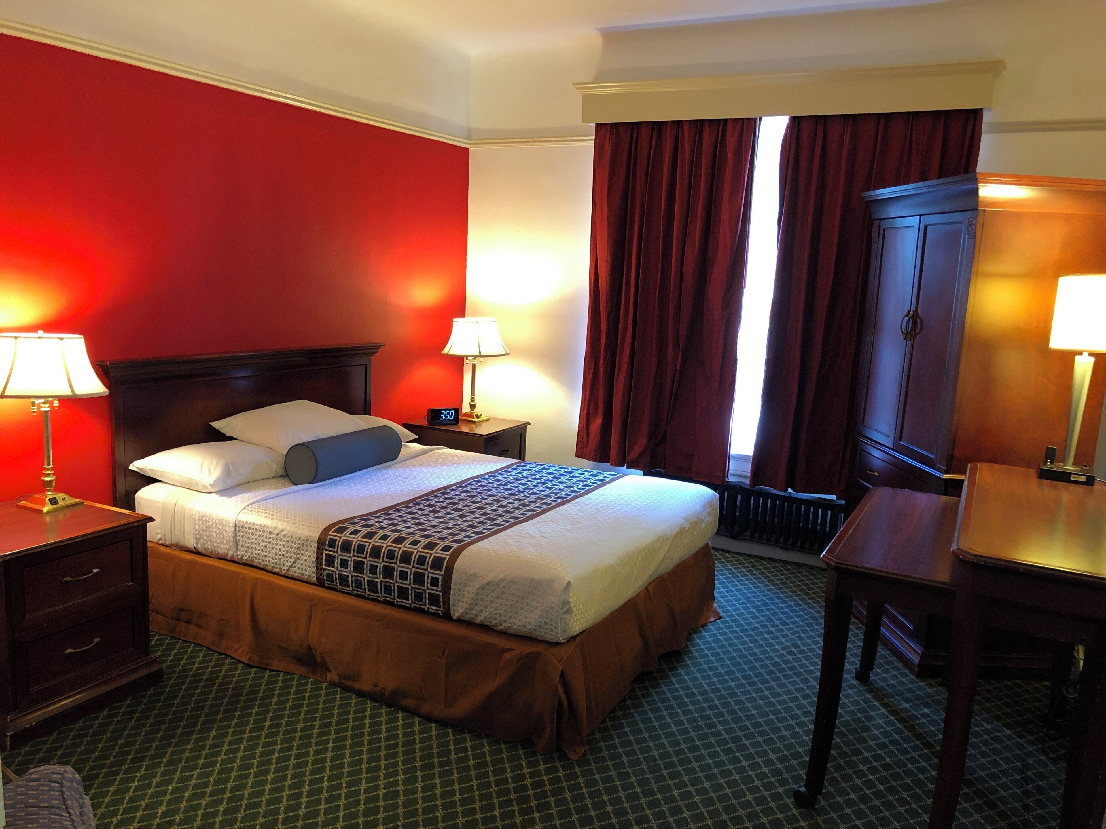 A hotel room with a bed, two bedside lamps, a nightstand, a wooden wardrobe, and a desk with a lamp.