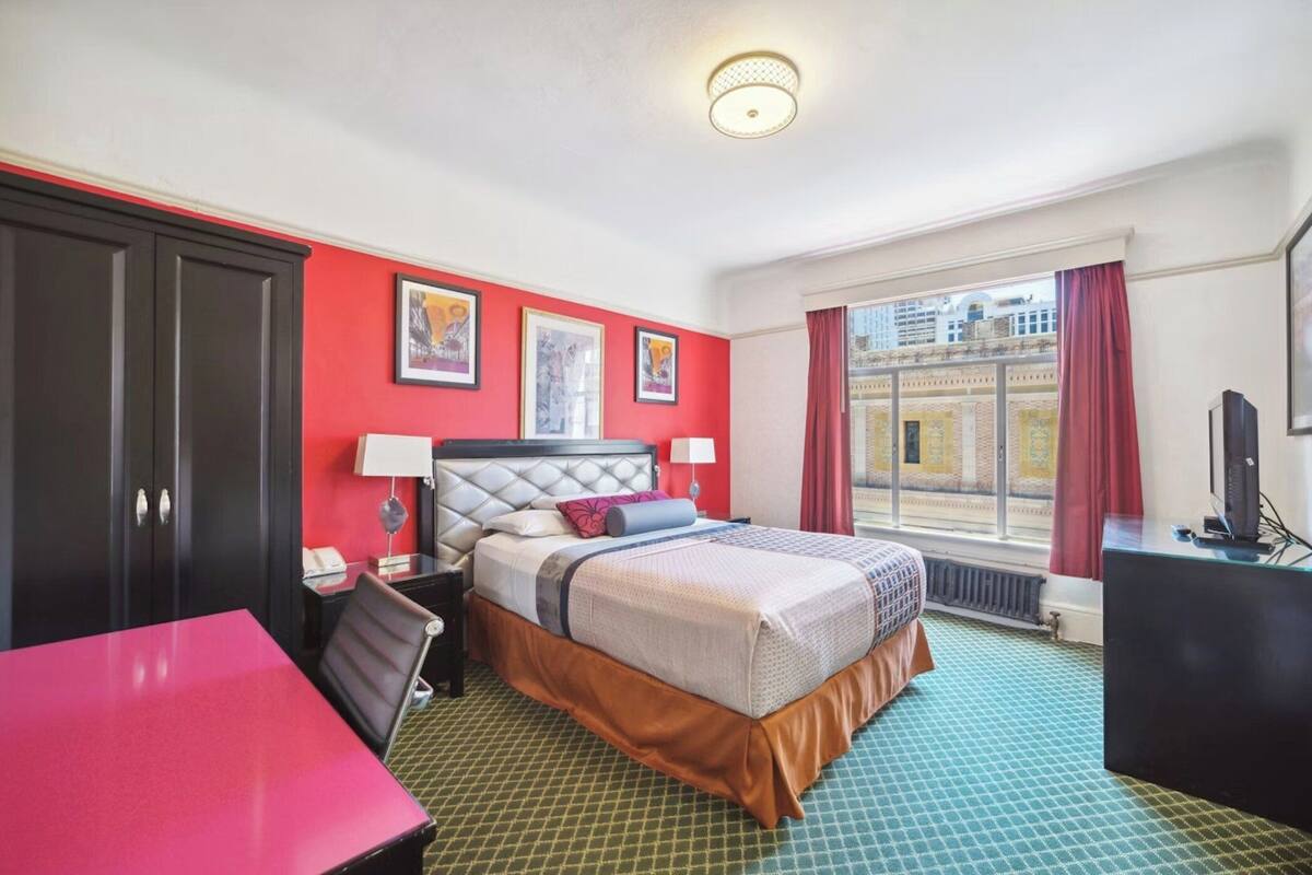 A hotel room with a bed, a desk, a chair, a TV, and a window with a view of buildings.