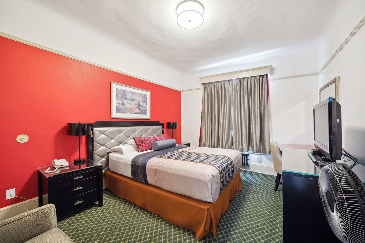 A hotel room with a red accent wall, a bed with a white headboard, a bedside table with a lamp, a TV, and a chair.