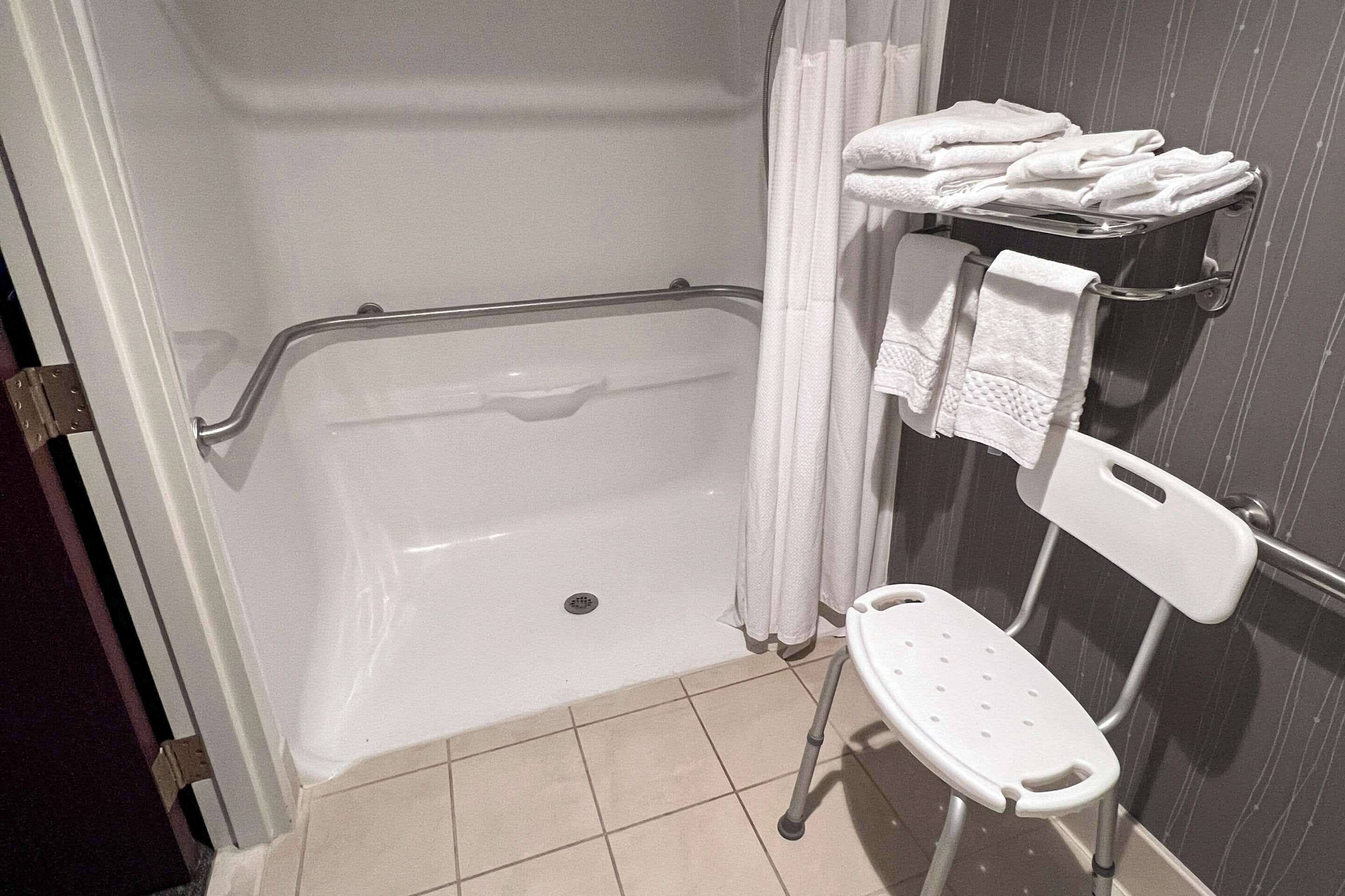 A bathroom with a walk-in shower, grab bars, a white chair, and a towel rack with folded towels.