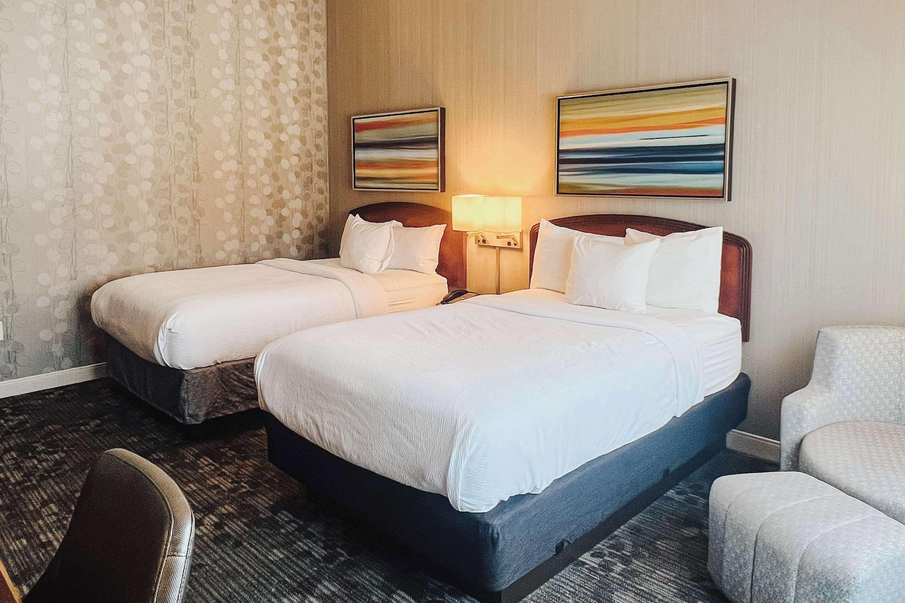 A hotel room with two beds, a desk, a chair, and two wall-mounted lamps.