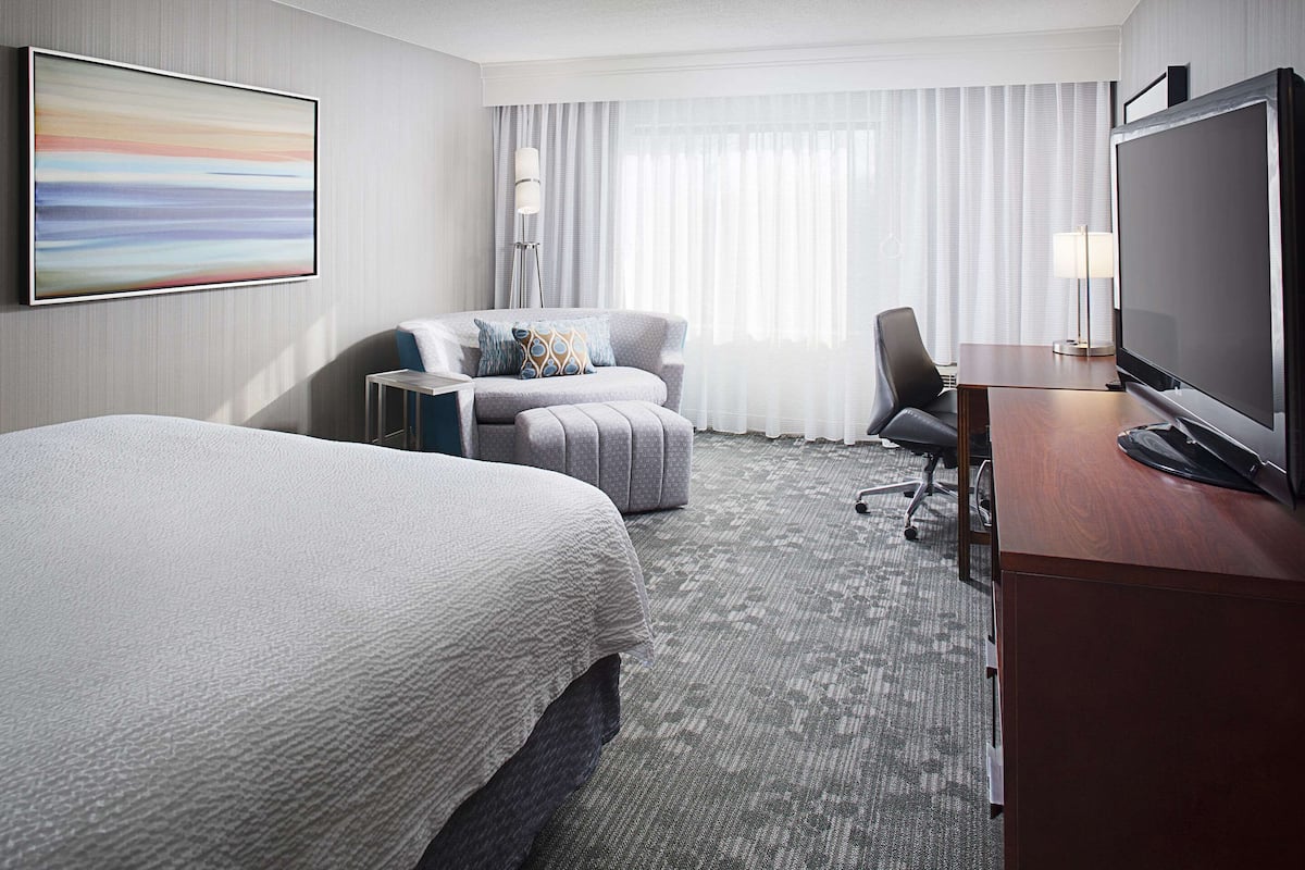 A hotel room with a bed, a desk with a TV, a chair, a sofa, and a large window with curtains.