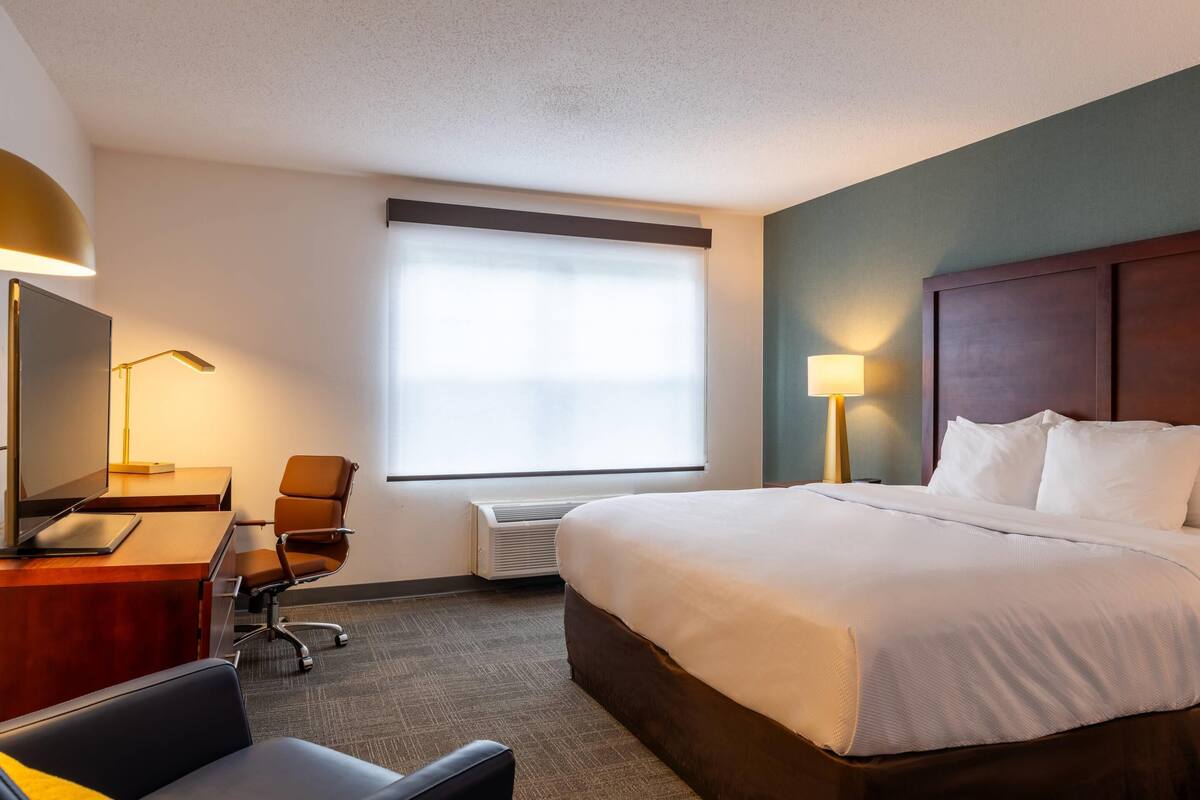 A hotel room with a bed, desk, chair, and a large window with blinds.