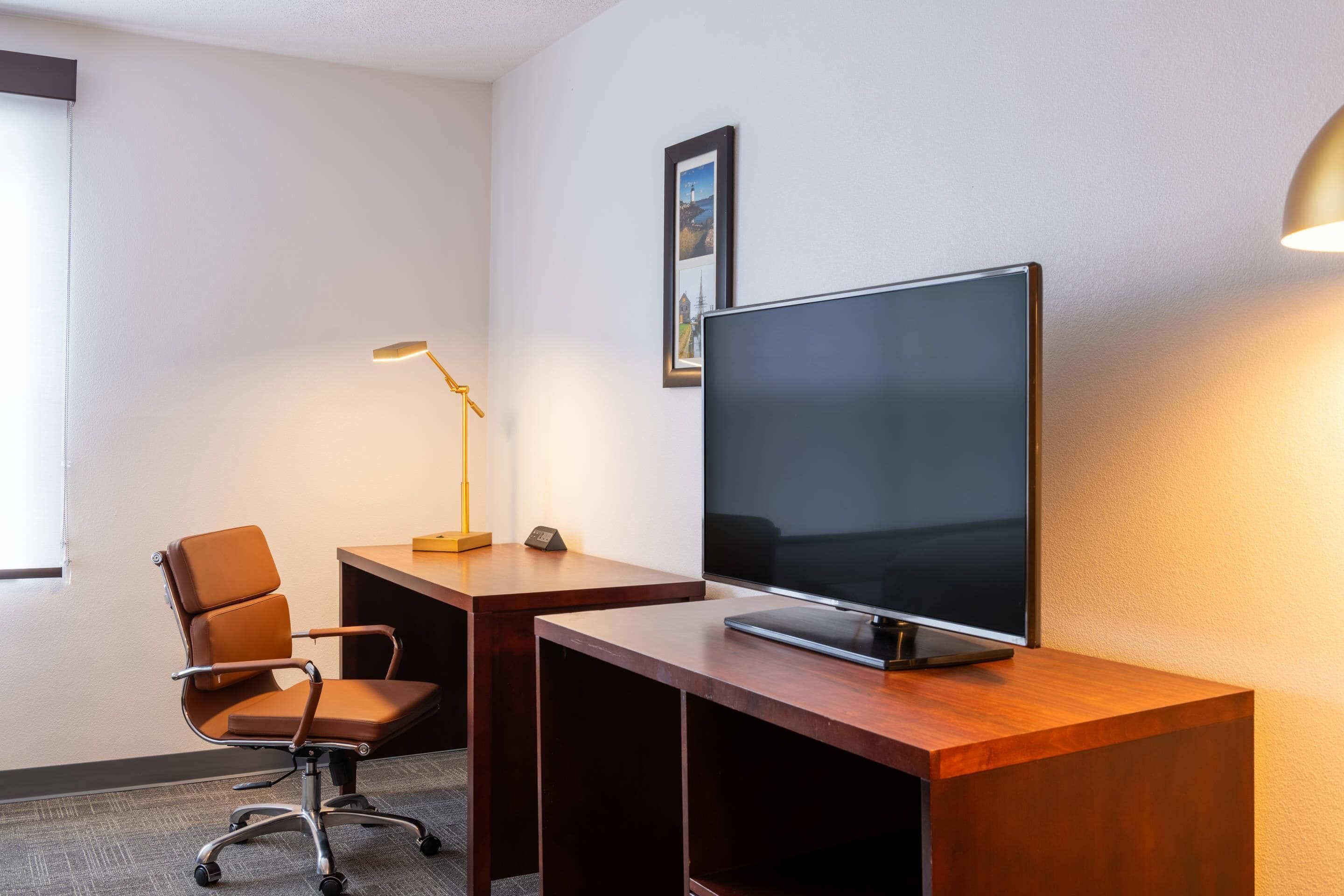 Habitación de hotel con un escritorio de madera, una silla de oficina marrón, un televisor de pantalla plana, una lámpara de escritorio y un cuadro en la pared.