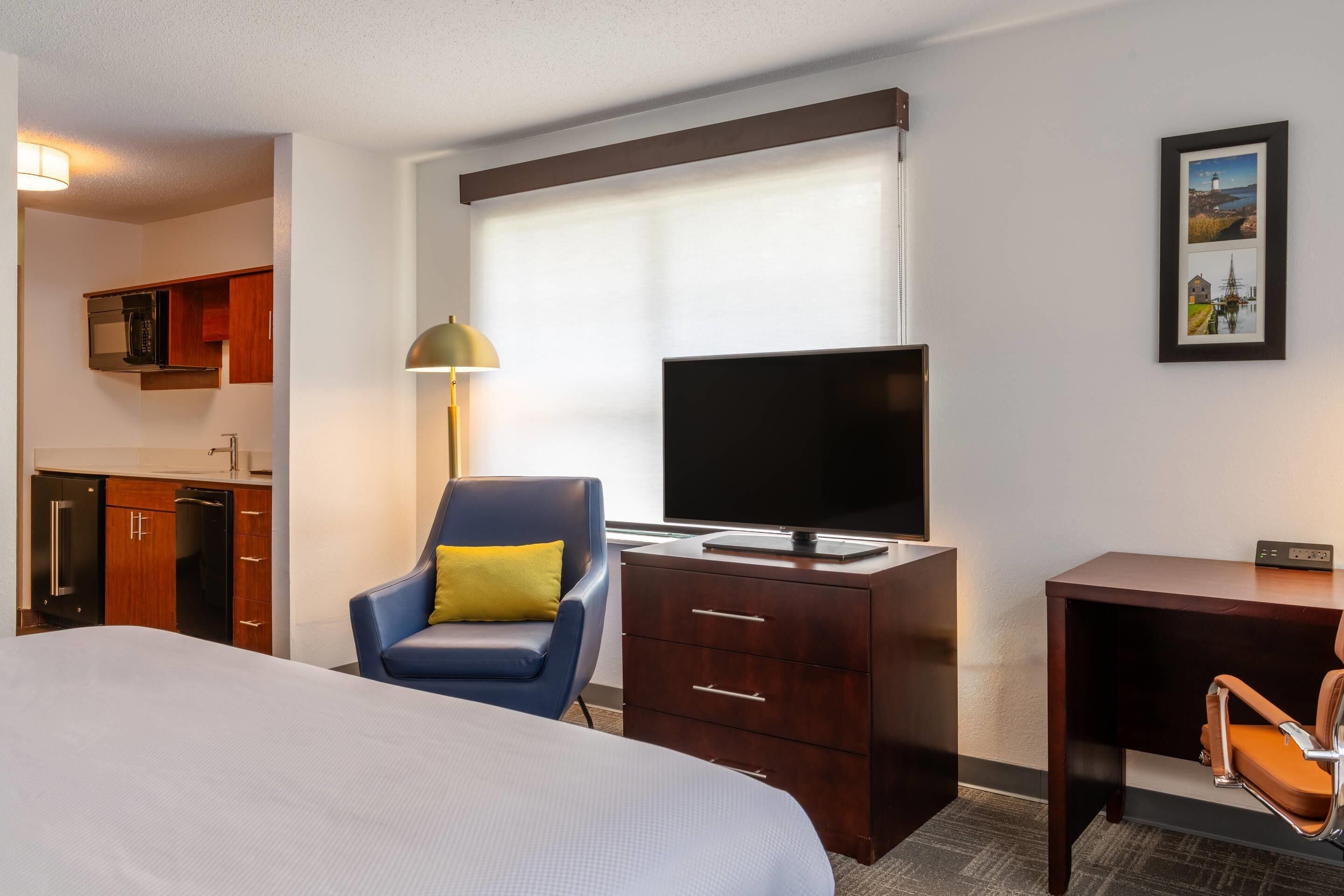 Habitación de hotel con cama, escritorio con televisor, silla, lámpara y un cuadro en la pared.