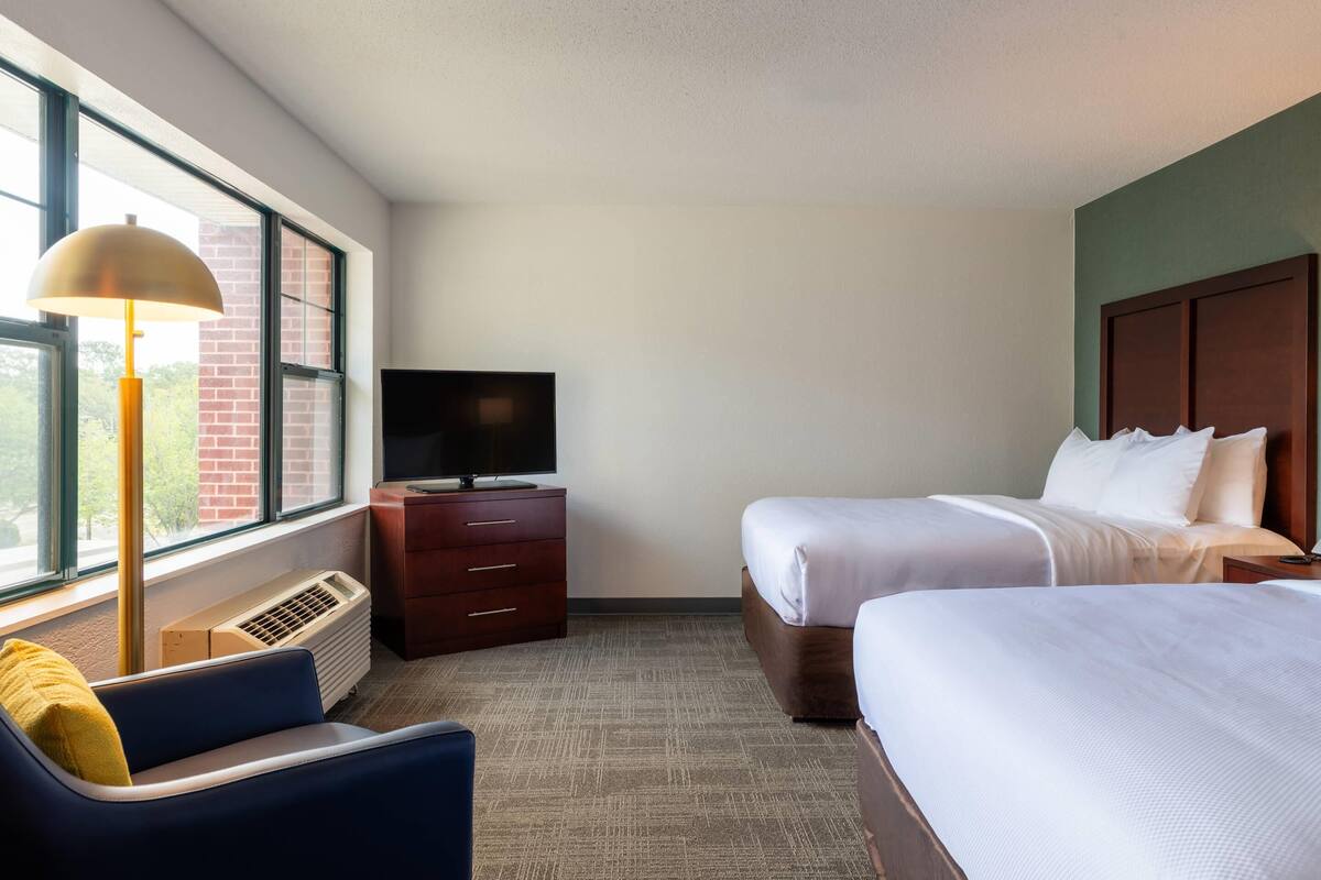A hotel room with two beds, a TV, a desk, and a window with a view of a brick building.