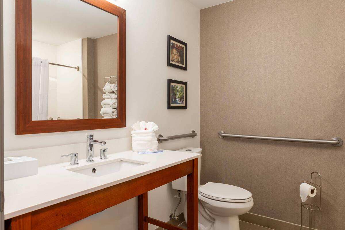 A bathroom with a white sink, mirror, and toilet. There are two framed pictures on the wall, a towel rack with towels, and a handrail.
